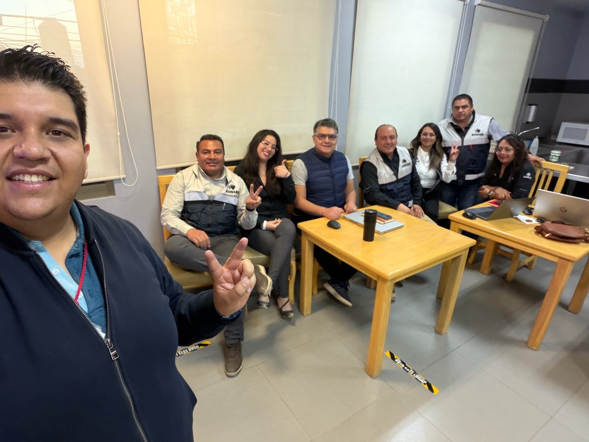 Hombre tomándose selfie con grupo de compañeros de trabajo sentados en mesas en sala de descanso de oficina