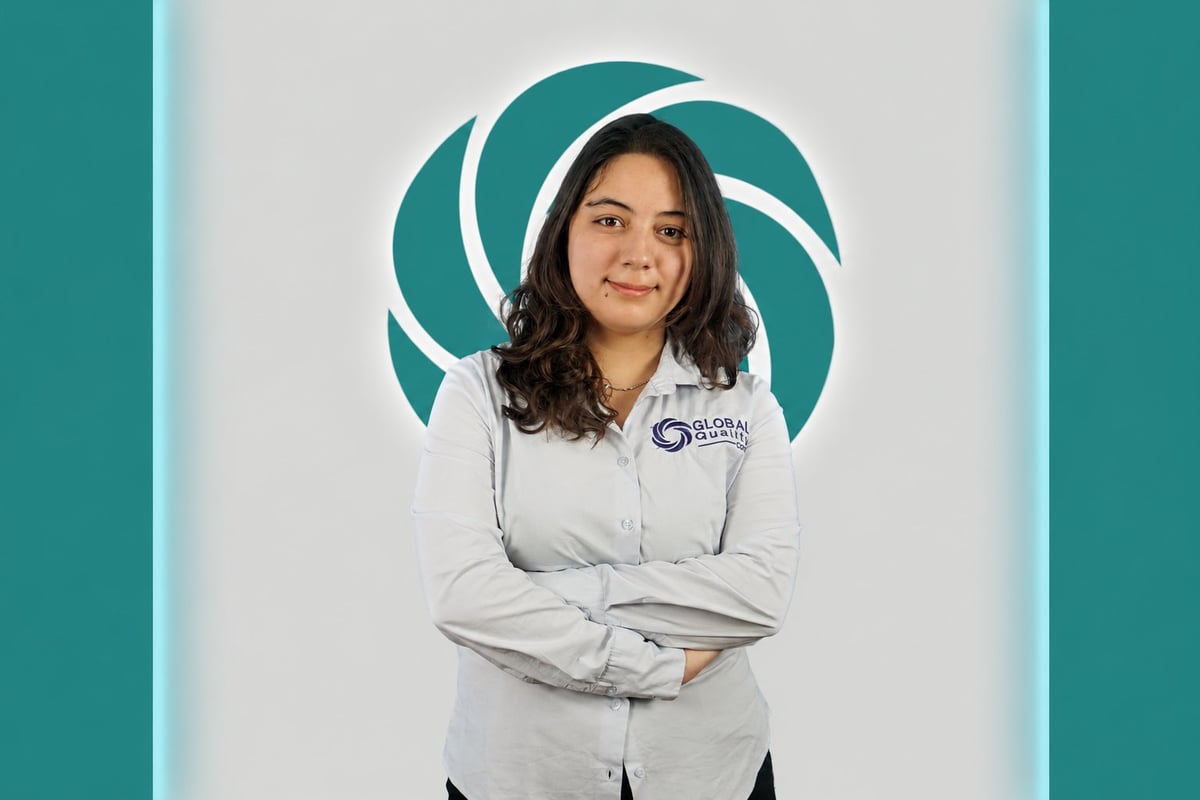 Professional headshot of a woman with long dark hair wearing a light gray jacket with arms crossed, against a teal and white circular background logo
